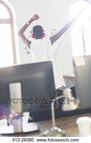 Stock Image - Casual businesswoman wearing headphones dancing in office. Fotosearch