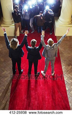 Stock Photo - Celebrities holding hands with arms raised for paparazzi photographers at red carpet event. Fotosearch