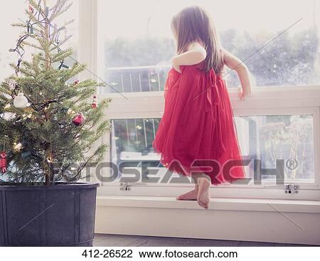 Stock Image - Girl in red dress on ledge next to potted Christmas tree. Fotosearch
