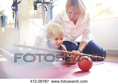 Stock Photo - Physical therapist guiding boy rolling ball with stick. Fotosearch