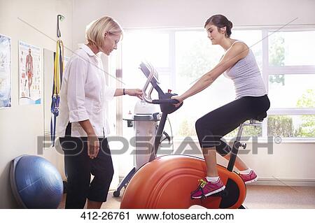 Stock Image - Physical therapist guiding woman on stationary bike. Fotosearch