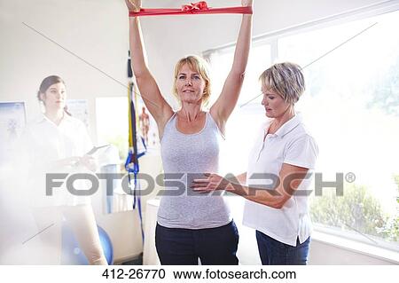 Stock Image - Physical therapist guiding woman pulling resistance band overhead. Fotosearch