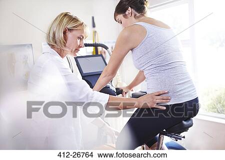 Picture - Physical therapist guiding woman’s hips on stationary bike. Fotosearch