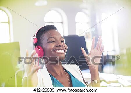 Stock Photo - Smiling businesswoman listening to music on headphones in office with eyes closed. Fotosearch