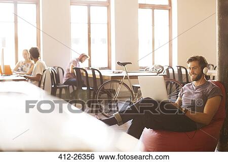 Stock Photograph - Smiling casual businessman wearing headphones and working at laptop on bean bag chair in office. Fotosearch