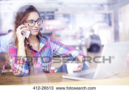 Stock Image - Woman using laptop and talking on cell phone at cafe. Fotosearch