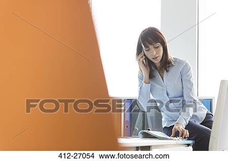 Picture - Brunette businesswoman talking on cell phone reviewing report in office. Fotosearch