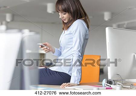 Stock Photograph - Brunette businesswoman texting with cell phone on desk in office. Fotosearch
