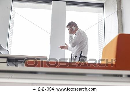 Businessman talking on cell phone gesturing at office window View Large Photo Image Picture - Businessman talking on cell phone gesturing at office window. Fotosearch