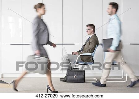 Businessman working at laptop in lobby behind business people on the move View Large Photo Image Stock Photo - Businessman working at laptop in lobby behind business people on the move. Fotosearch