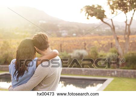 Stock Image - Couple hugging and looking at mountain view poolside. Fotosearch