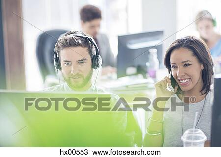 Stock Image - Creative business people with headphones talking on cell phone and using computer in office. Fotosearch