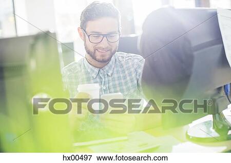 Stock Photo - Creative businessman drinking coffee and texting with cell phone at computer. Fotosearch