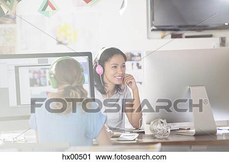 Stock Image - Creative businesswomen listening to headphones and working at computers in office. Fotosearch