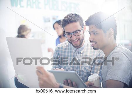 Stock Photograph - Creative editors with coffee using digital tablet and reviewing paperwork. Fotosearch