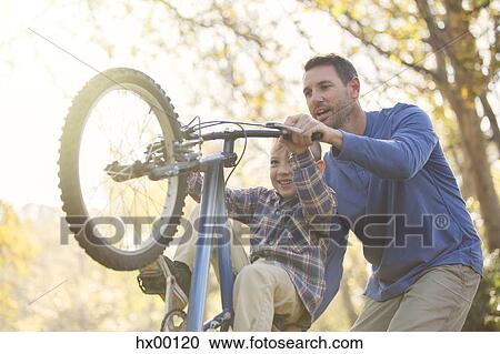 Stock Image - Father teaching son wheelie on bicycle. Fotosearch