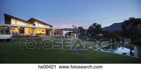 Stock Image - Illuminated modern house beyond yard and swimming pool at night. Fotosearch
