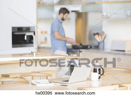 Stock Photography - Man cooking at stove behind laptop, coffee and cell phone in kitchen. Fotosearch