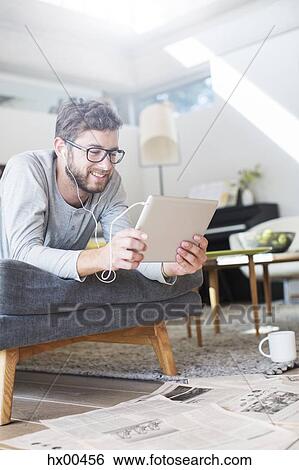Stock Photograph - Man laying with headphones and using digital tablet in living room. Fotosearch