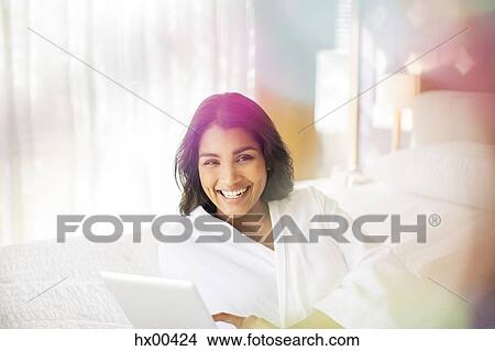 Picture - Portrait smiling woman in bathrobe using digital tablet on bed. Fotosearch