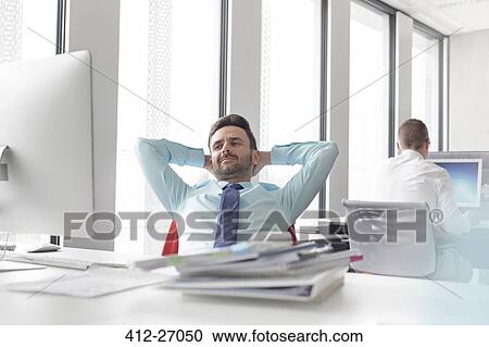 Stock Image - Relaxed businessman leaning with hands behind head at computer in office. Fotosearch