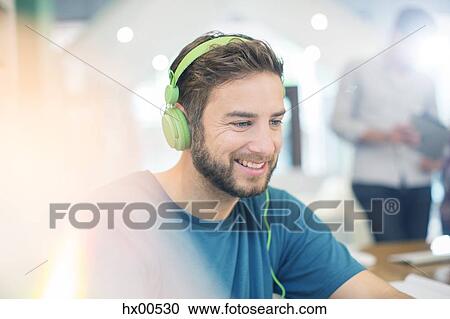 Stock Image - Smiling creative businessman listening to headphones in office. Fotosearch