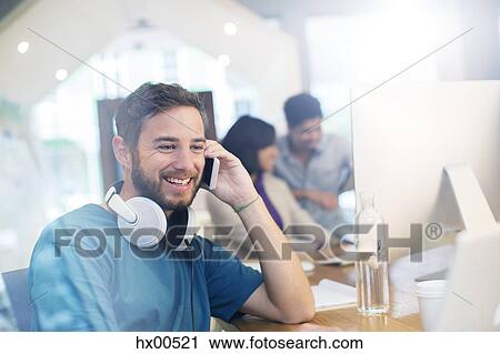 Stock Image - Smiling creative businessman with headphones talking on cell phone in office. Fotosearch