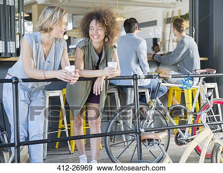 Stock Photo - Smiling friends hanging out drinking coffee at cafe patio railing near bicycles. Fotosearch