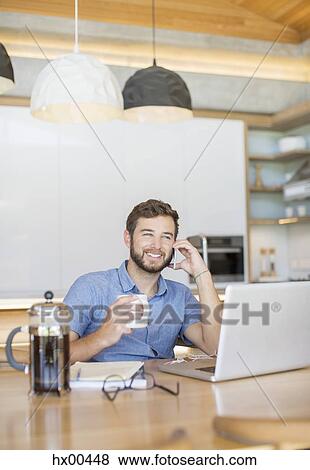 Stock Photo - Smiling man drinking coffee and talking on cell phone at laptop. Fotosearch