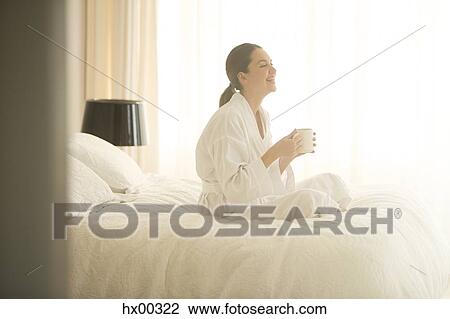 Stock Image - Smiling woman in bathrobe drinking coffee cross-legged on bed. Fotosearch