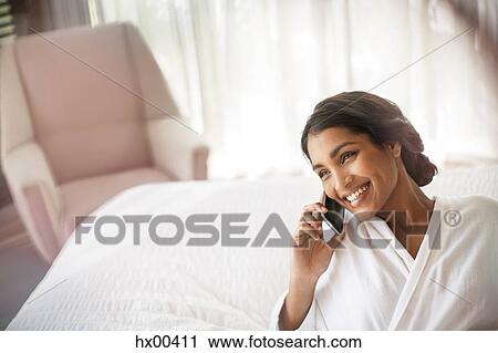 Stock Image - Smiling woman in bathrobe talking on cell phone on bed. Fotosearch