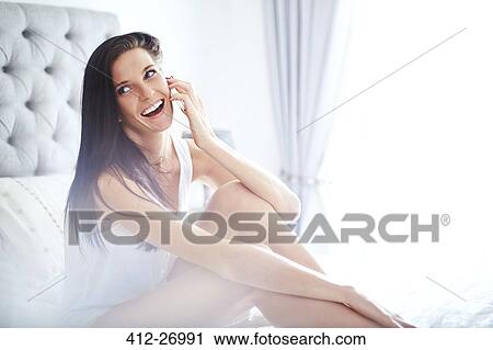 Stock Image - Smiling woman talking on cell phone on bed. Fotosearch