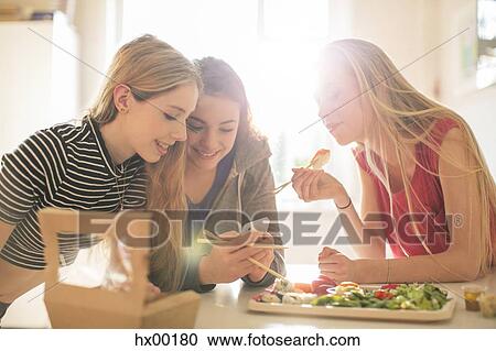 Stock Image - Teenage girls eating sushi and texting with cell phone in sunny kitchen. Fotosearch