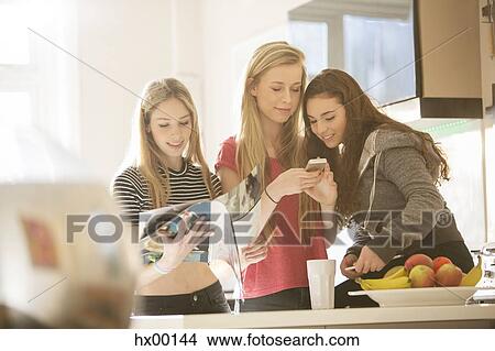 Picture - Teenage girls reading magazine and using cell phone in kitchen. Fotosearch