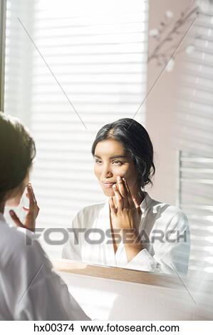 Picture - Woman applying moisturizer to face in bathroom mirror. Fotosearch