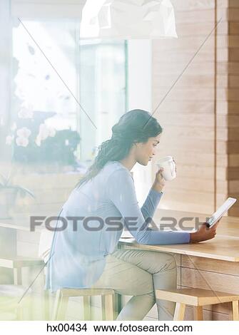 Picture - Woman drinking coffee and using digital tablet at breakfast bar. Fotosearch
