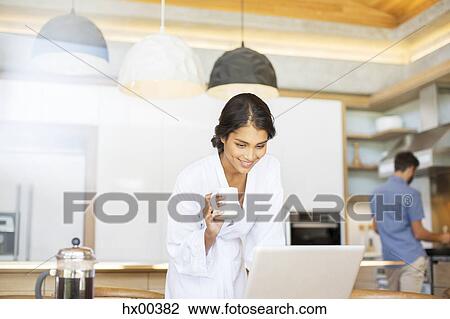 Stock Image - Woman in bathrobe drinking coffee and using laptop in kitchen. Fotosearch