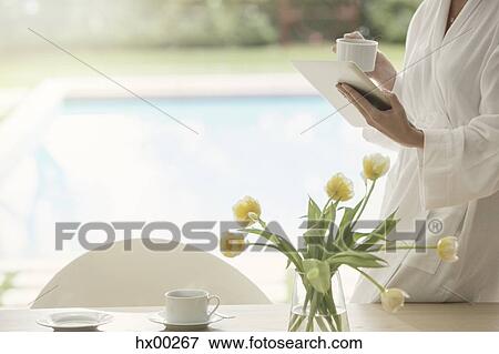Stock Photo - Woman in bathrobe drinking coffee and using digital tablet at poolside. Fotosearch