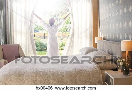 Stock Photograph - Woman in bathrobe opening bedroom curtains. Fotosearch