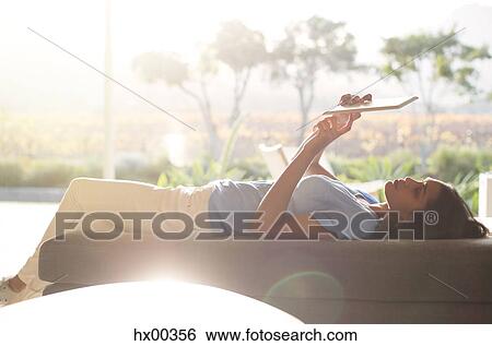 Stock Photograph - Woman laying on chaise lounge using digital tablet on sunny patio. Fotosearch