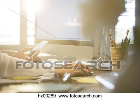 Stock Photo - Woman using computer and texting on cell phone at desk. Fotosearch