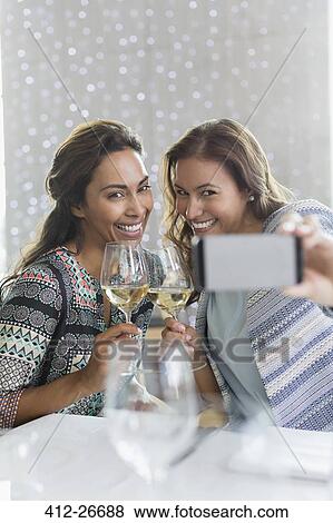Stock Photo - Women with white wine taking selfie with camera phone in restaurant. Fotosearch