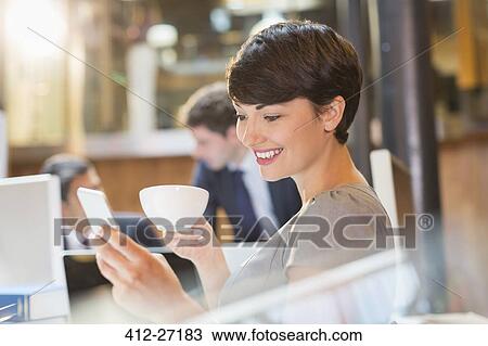 Stock Image - Businesswoman drinking coffee and texting with cell phone in office. Fotosearch