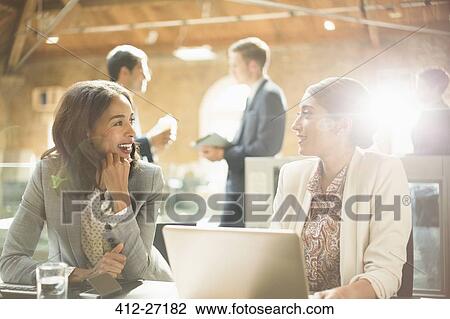 Stock Image - Businesswomen talking and working at laptop in office. Fotosearch