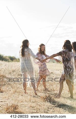 Stock Image - Boho women holding hands and dancing in circle in sunny rural field. Fotosearch