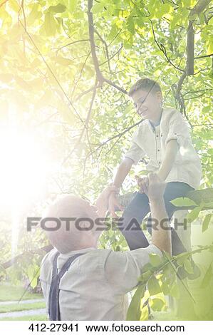 Stock Image - Grandfather helping grandson off sunny tree branch. Fotosearch