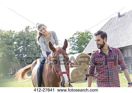 Stock Image - Man leading woman horseback riding in rural pasture. Fotosearch