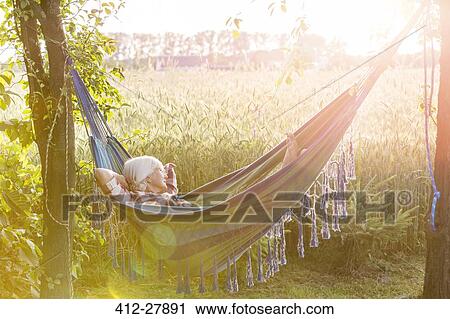 Stock Image - Serene woman napping in hammock next to sunny rural wheat field. Fotosearch