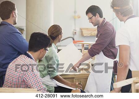 Stock Image - Carpentry teacher explaining blueprints to students in workshop. Fotosearch