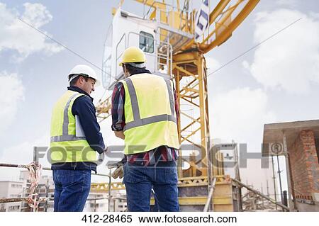 Stock Photography - Construction worker and engineer reviewing blueprints below crane at construction site. Fotosearch